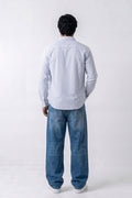 A man viewed from behind wearing a light blue long-sleeve button-up linen shirt and loose blue jeans against a plain white studio background.