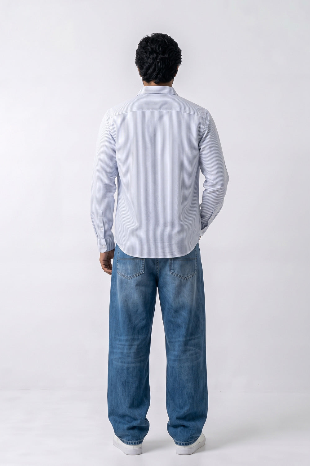 A man viewed from behind wearing a light blue long-sleeve button-up linen shirt and loose blue jeans against a plain white studio background.