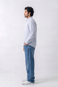A man standing in profile wearing a light blue long-sleeve button-up linen shirt, blue jeans, and white sneakers against a plain light gray studio background.