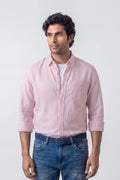 A man wearing a light pink and white vertically striped button-down linen shirt with rolled-up sleeves, paired with blue jeans and a dark blue patterned belt, standing against a light gray studio background.
