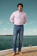 A man standing on a boat deck wearing a light pink long-sleeve button-down linen shirt and blue jeans, with water and sky in the background.