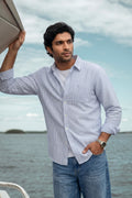 A man wearing a light blue and white vertically striped button-down linen shirt with blue jeans, standing on a boat with water and sky in the background.