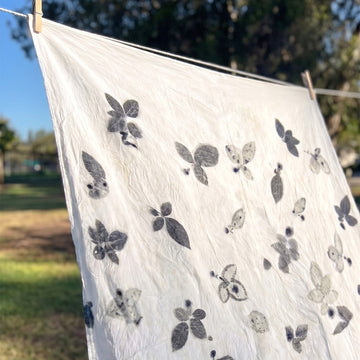A white fabric sheet or towel with black floral and leaf eco-prints hangs from a clothesline outdoors, secured with wooden clothespins against a blurred grassy background.