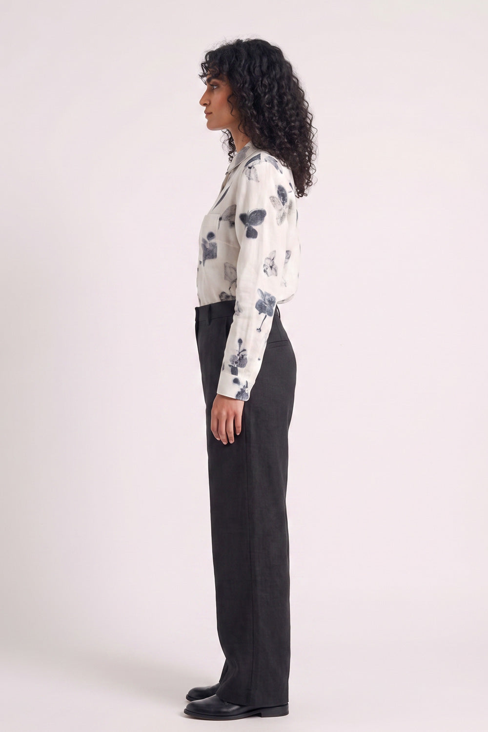 A model stands against a plain light background wearing a white long-sleeve button-up shirt with a gray eco-printed floral pattern, paired with black wide-leg trousers and black shoes.
