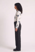 A woman stands in profile wearing a white long-sleeve blouse with a dark grey eco-printed floral print and black wide-leg trousers, photographed against a plain light background.