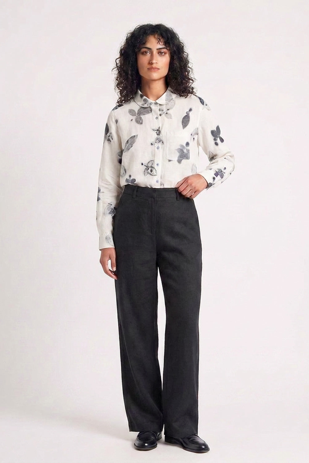 A model stands against a plain light background wearing a white long-sleeve button-up shirt with a gray eco-printed floral pattern, paired with black wide-leg trousers and black shoes.