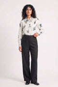 A model stands against a plain light background wearing a white long-sleeve button-up shirt with a gray eco-printed floral pattern, paired with black wide-leg trousers and black shoes.