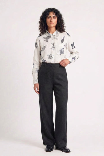 A model stands against a plain light background wearing a white long-sleeve button-up shirt with a gray eco-printed floral pattern, paired with black wide-leg trousers and black shoes.