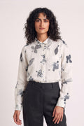A woman with curly dark hair wearing a white long-sleeve button-up shirt with a gray eco-print floral and dragonfly print, paired with dark charcoal trousers, against a light off-white studio background.