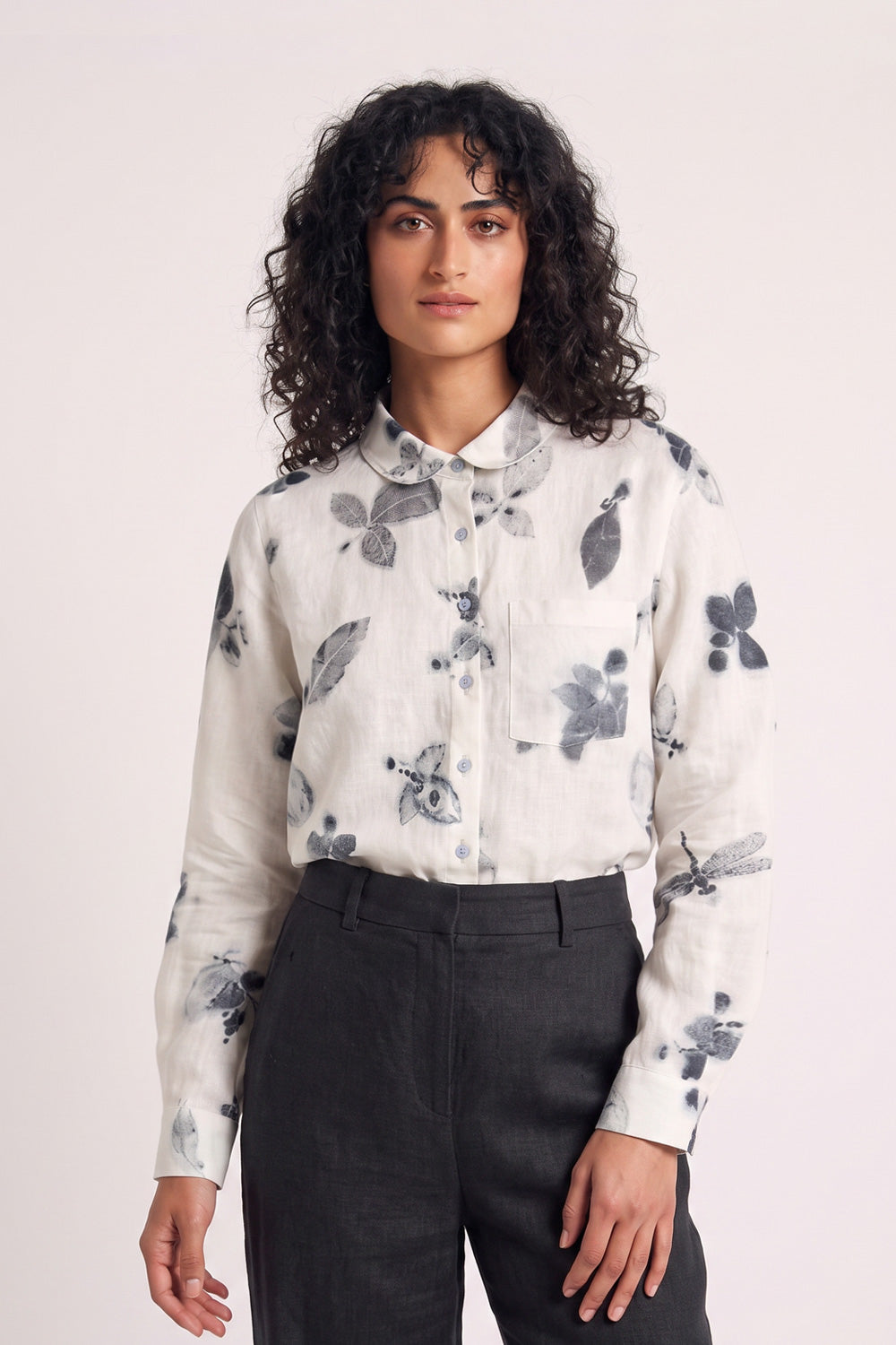 A woman with curly dark hair wearing a white long-sleeve button-up shirt with a gray eco-print floral and dragonfly print, paired with dark charcoal trousers, against a light off-white studio background.