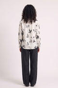 A model shown from the back wearing a cream-colored long-sleeve blouse with a black and gray eco-printed print, paired with black wide-leg trousers, against a plain light background.