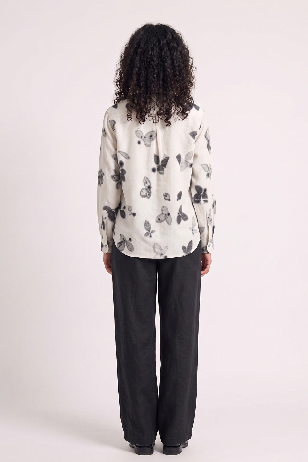 A model shown from the back wearing a cream-colored long-sleeve blouse with a black and gray eco-printed print, paired with black wide-leg trousers, against a plain light background.