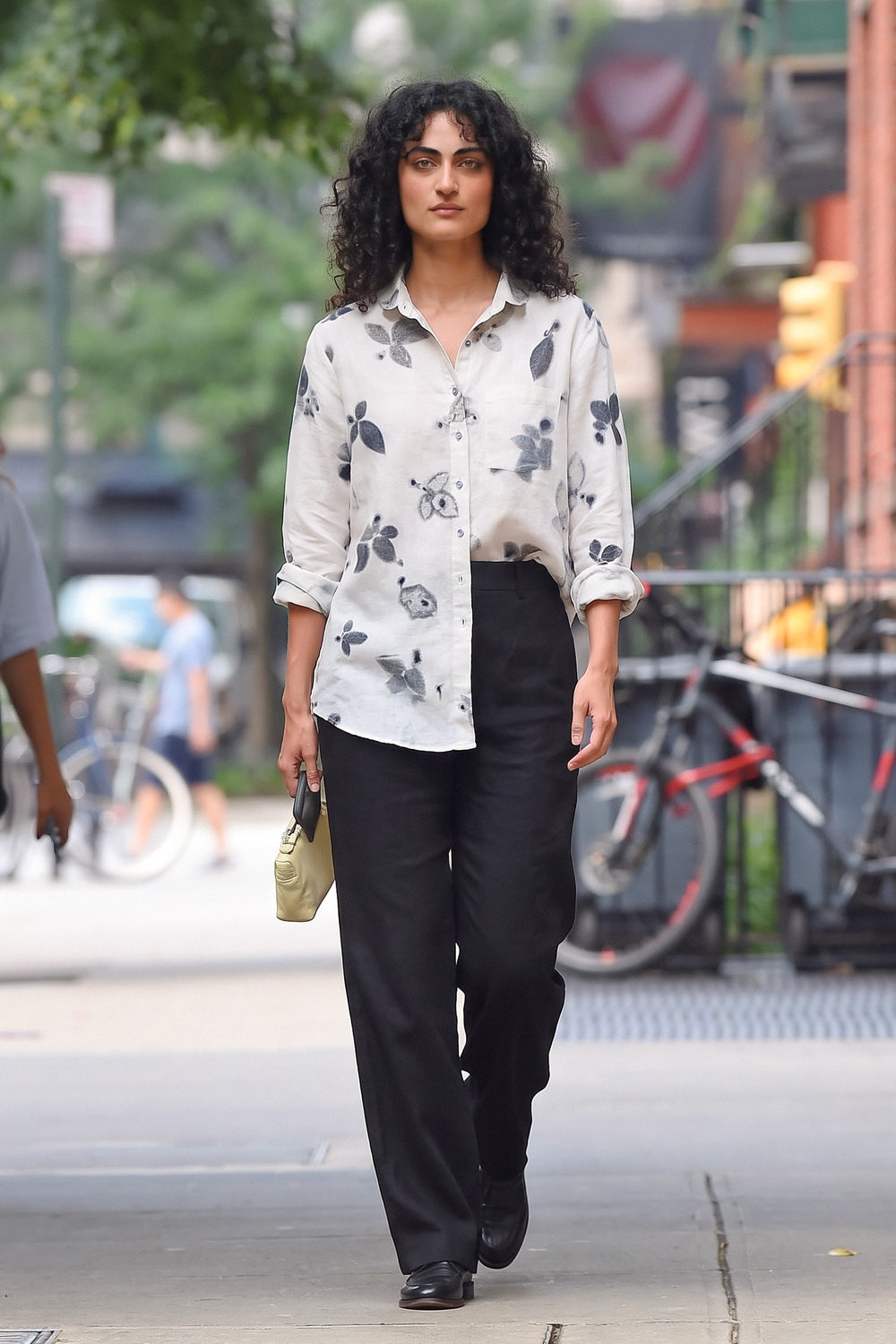 A woman walking on a city sidewalk wearing a white long-sleeve button-up shirt with a black eco-print floral print, black wide-leg trousers, and carrying a small beige handbag.