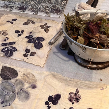 Several cream-colored fabric sheets laid out on a table, each printed with dark botanical leaf and flower stencils, alongside a metal colander filled with dried leaves and herbs.