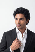 A man with dark curly hair wearing a black hemp suit jacket over a white button-down shirt, adjusting his collar against a plain white background.