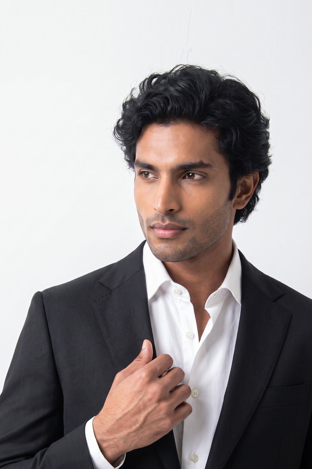 A man with dark curly hair wearing a black hemp suit jacket over a white button-down shirt, adjusting his collar against a plain white background.