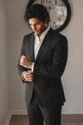A man wearing a black hemp suit jacket and trousers over a white dress shirt, adjusting his cuff while standing in an indoor setting with a wall clock in the background.
