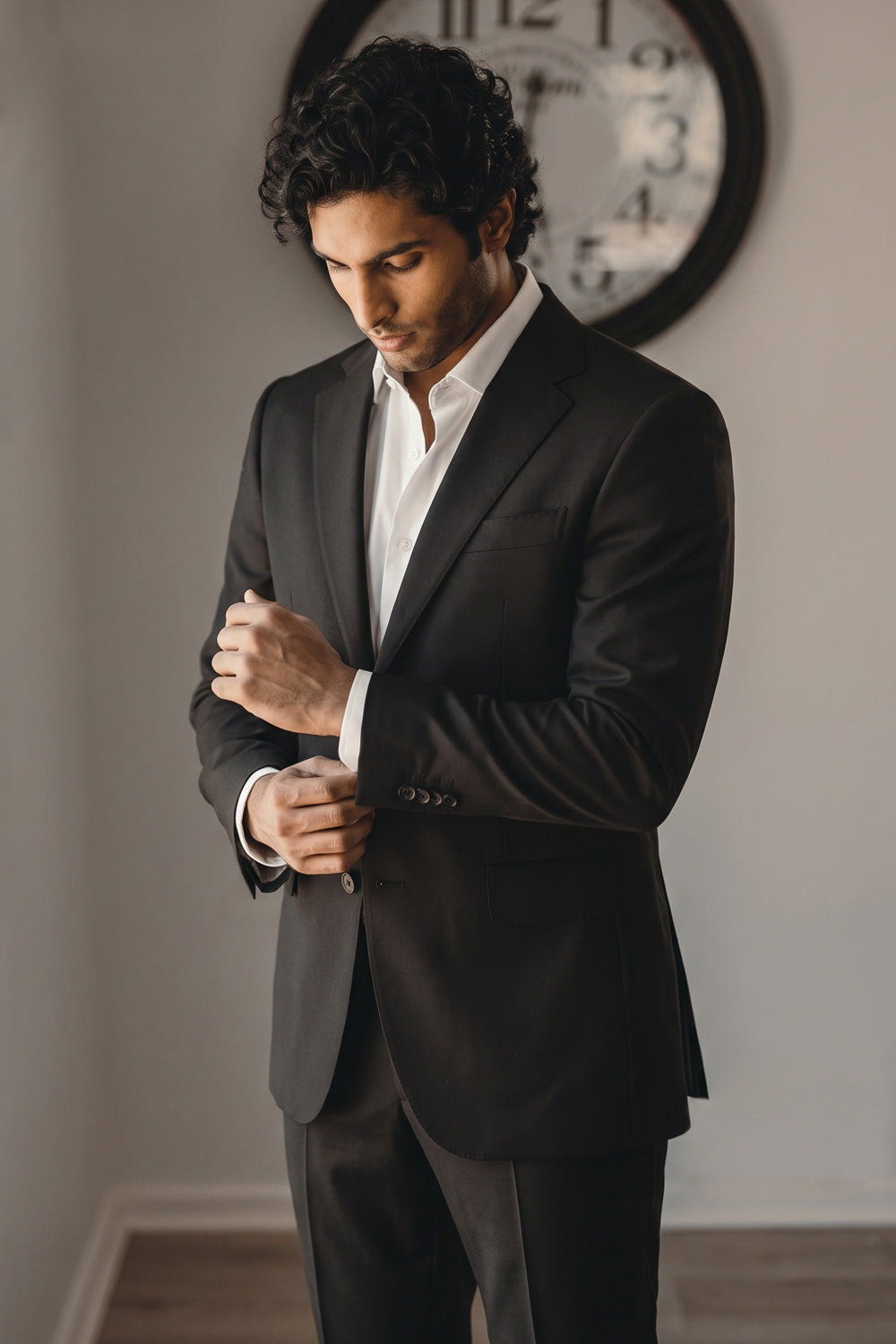 A man wearing a black hemp suit jacket and trousers over a white dress shirt, adjusting his cuff while standing in an indoor setting with a wall clock in the background.