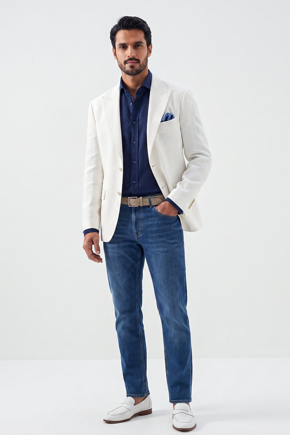 A man modeling a white hemp blazer over a navy blue button-down shirt, paired with blue jeans, a beige belt, and white loafers, standing against a plain white studio background.
