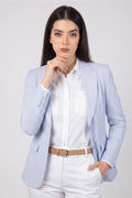 A woman modeling a light blue and white pinstriped linen blazer over a white button-down shirt and white trousers, accessorized with a tan belt, against a light gray studio background.