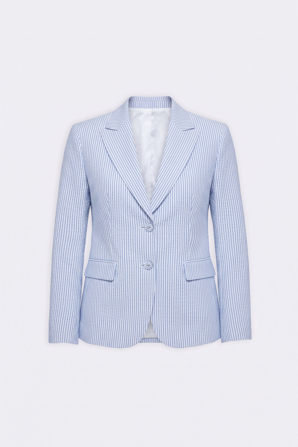 A woman modeling a light blue linen blazer over a white shirt and white trousers, styled with a thin brown belt and white heels, against a plain light gray studio background.