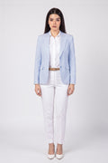 A woman modeling a light blue linen blazer over a white shirt and white trousers, styled with a thin brown belt and white heels, against a plain light gray studio background.