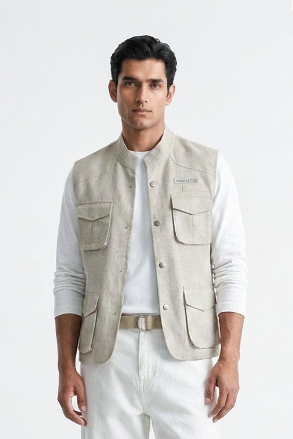 A man modeling a light beige utility linen vest over a white long-sleeve T-shirt, paired with white trousers, a beige belt, and white sneakers, against a plain white studio background.