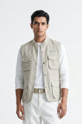 A man wearing a light beige linen utility linen vest over a white long-sleeve T-shirt and white trousers, standing against a plain white studio background.