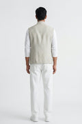 A man viewed from the back wearing a light beige linen vest over a white long-sleeve shirt and white trousers, standing against a plain white studio background.
