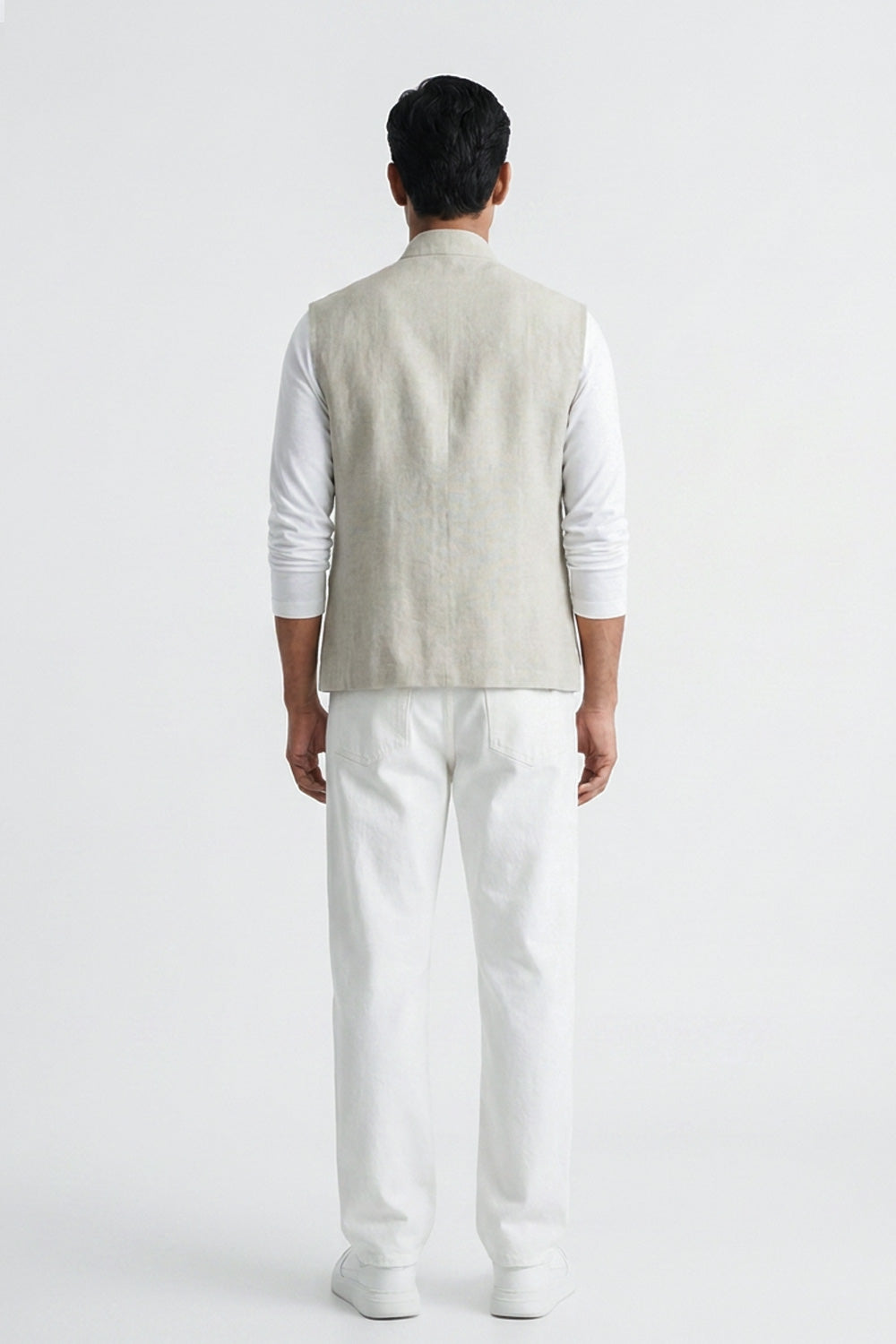 A man viewed from the back wearing a light beige linen vest over a white long-sleeve shirt and white trousers, standing against a plain white studio background.