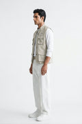 A man wearing a light beige linen utility linen vest over a white long-sleeve T-shirt and white trousers, standing in profile against a plain white studio background.