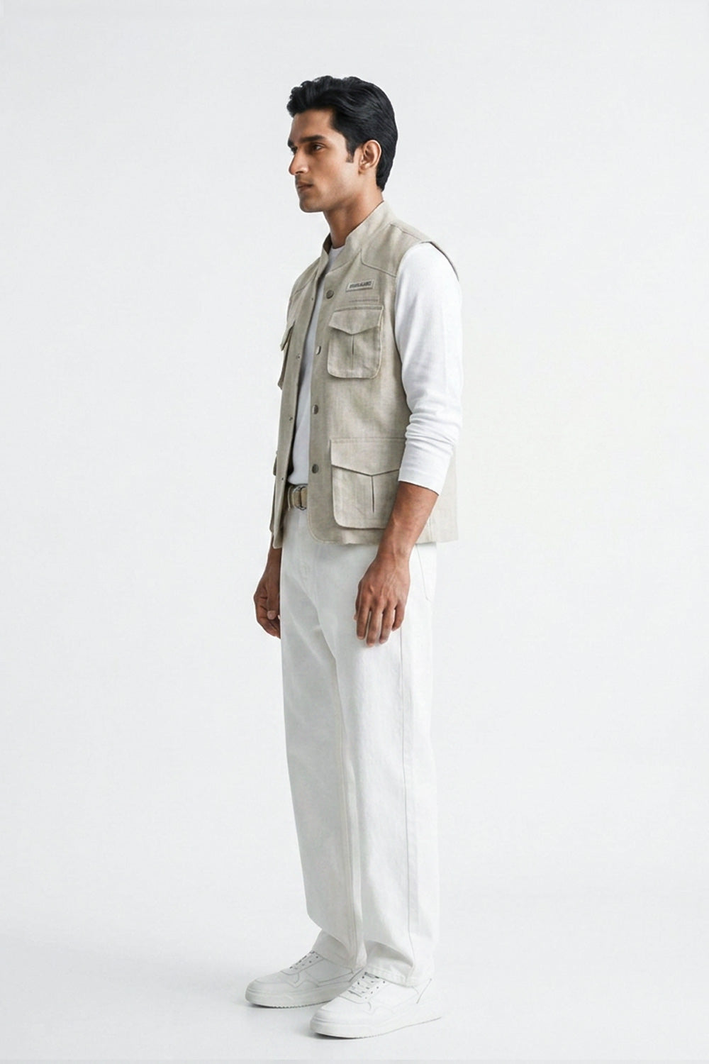 A man wearing a light beige linen utility linen vest over a white long-sleeve T-shirt and white trousers, standing in profile against a plain white studio background.