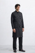 A man modeling a dark charcoal gray traditional Indian outfit consisting of a sleeve-less button-up linen jacket with multiple chest pockets, a matching long kurta, trousers, and dark loafers, set against a plain light gray studio background.
