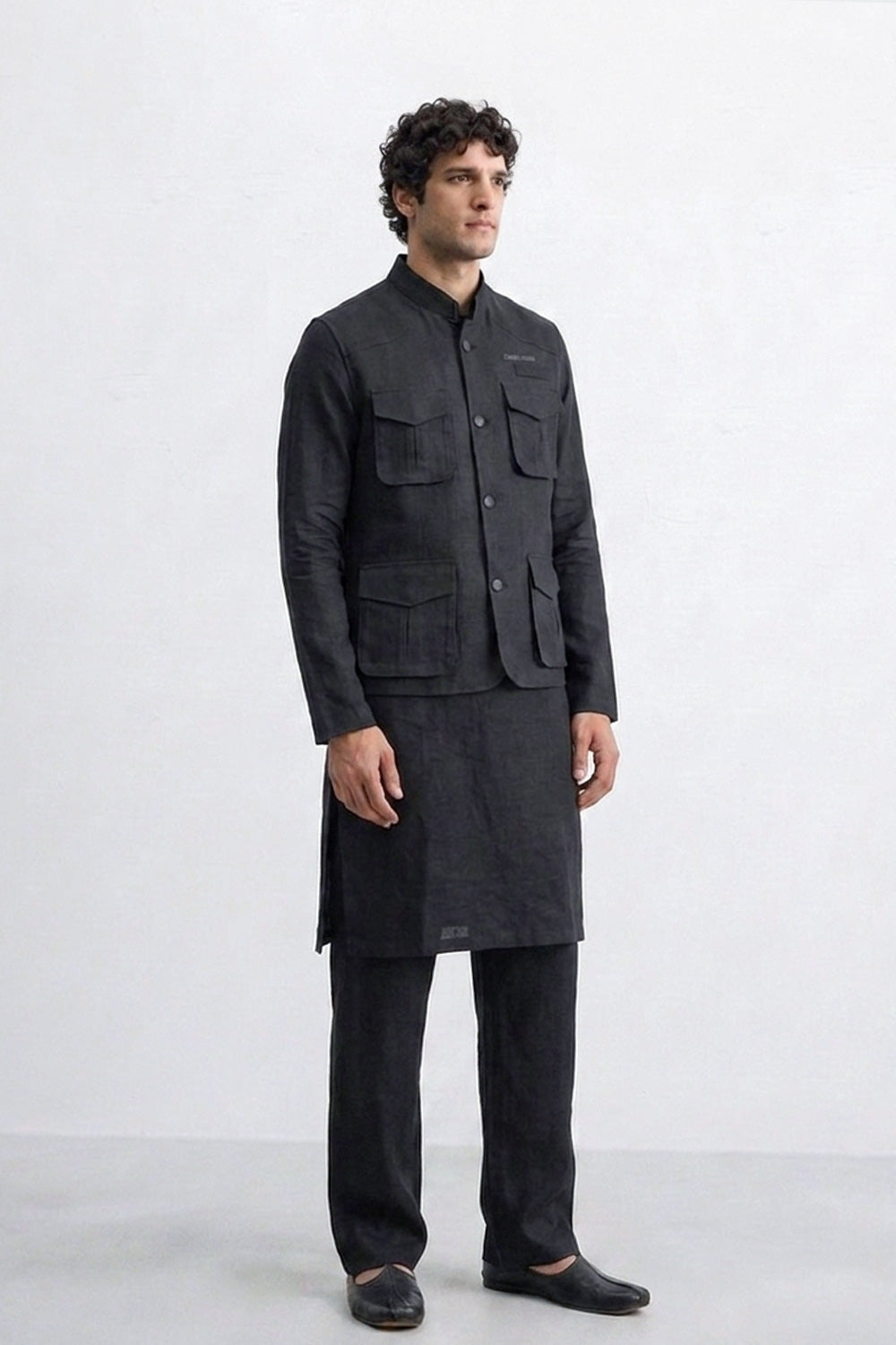 A man modeling a dark charcoal gray traditional Indian outfit consisting of a sleeve-less button-up linen jacket with multiple chest pockets, a matching long kurta, trousers, and dark loafers, set against a plain light gray studio background.