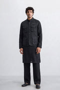 A man modeling a black traditional outfit consisting of a sleeve-less linen jacket with multiple front pockets, a matching long kurta, wide-leg trousers, and black shoes, set against a plain light gray studio background.