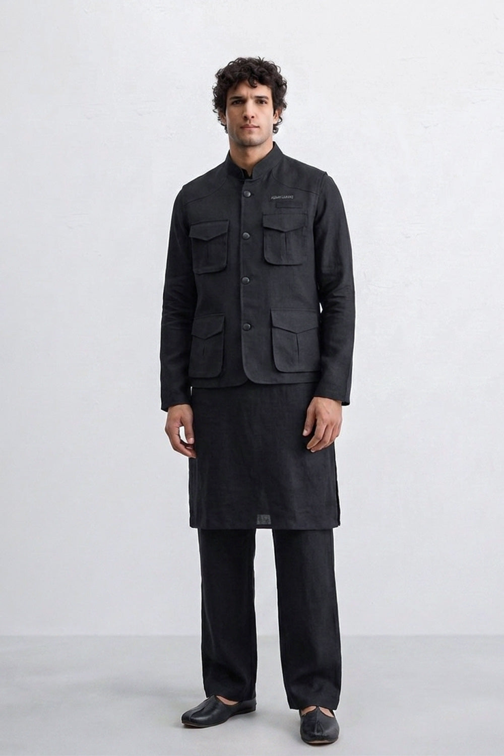 A man modeling a black traditional outfit consisting of a sleeve-less linen jacket with multiple front pockets, a matching long kurta, wide-leg trousers, and black shoes, set against a plain light gray studio background.