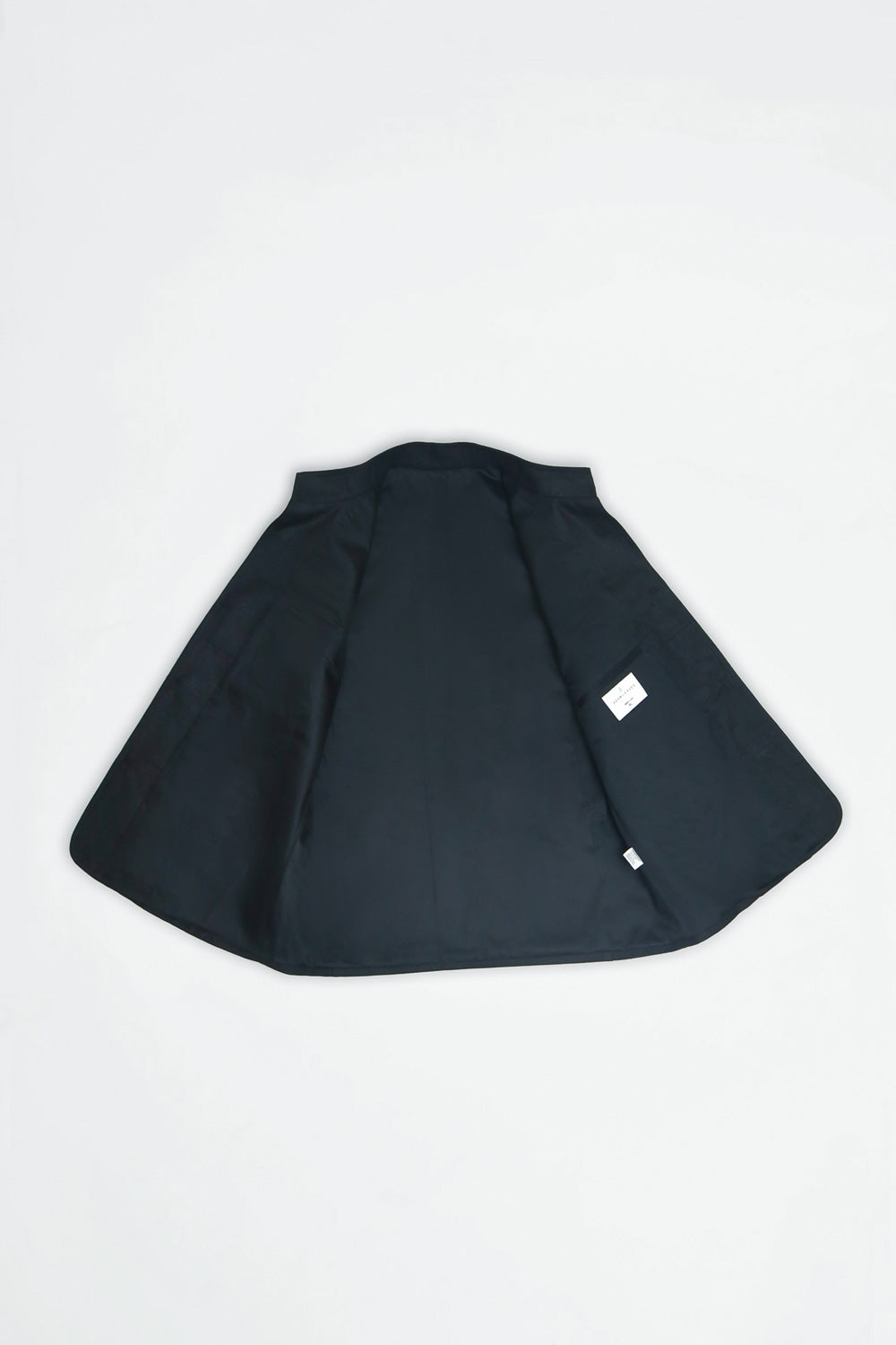 A black inside of jacket or cape-style outerwear piece laid flat on a light gray background, featuring a small white rectangular tag near the lower right side.