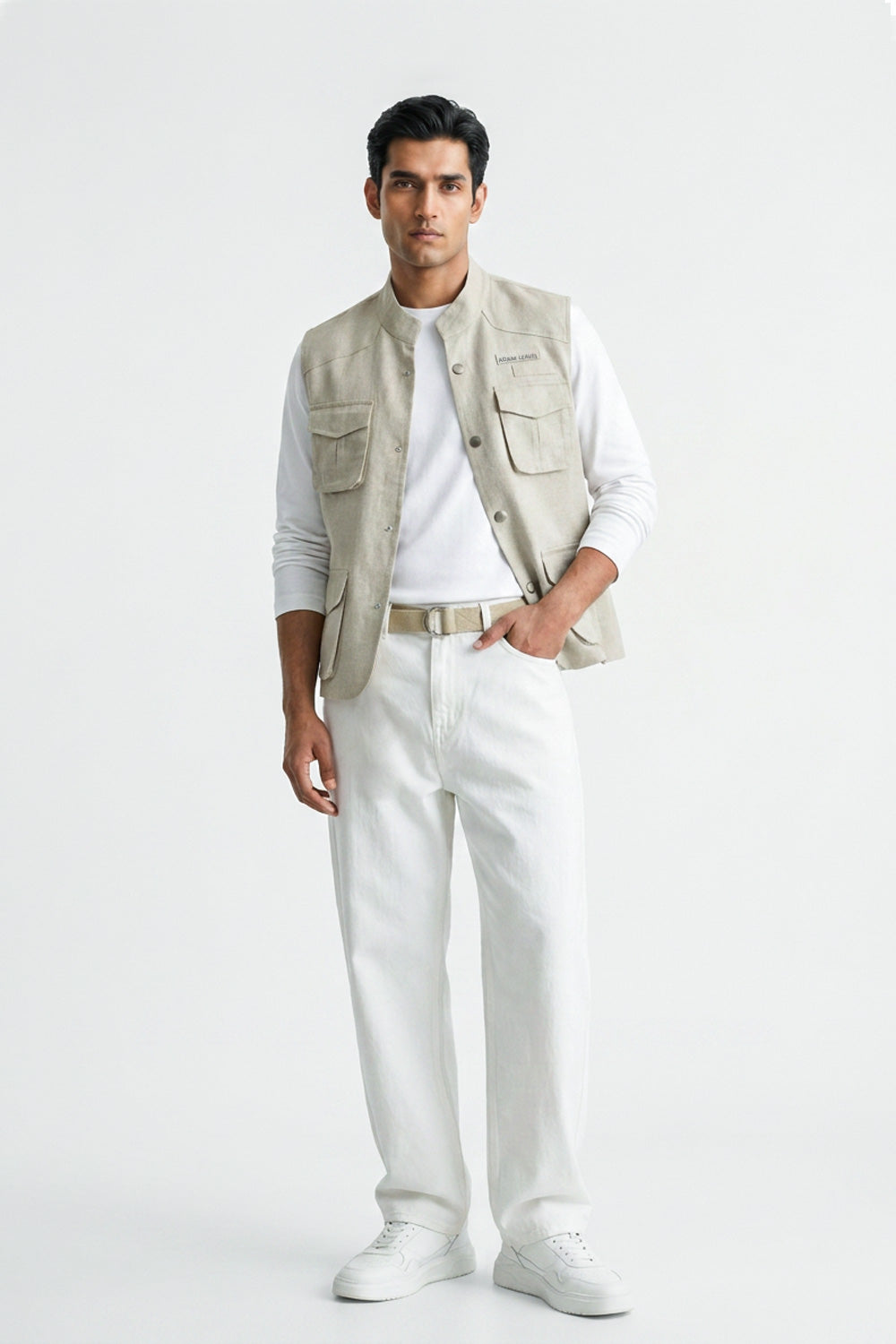 A man modeling a light beige utility linen vest over a white long-sleeve T-shirt, paired with white trousers, a beige belt, and white sneakers, against a plain white studio background.