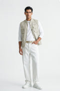 A man modeling a light beige utility linen vest over a white long-sleeve T-shirt, paired with white trousers, a beige belt, and white sneakers, against a plain white studio background.