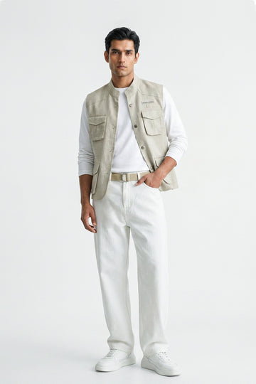 A man modeling a light beige utility linen vest over a white long-sleeve T-shirt, paired with white trousers, a beige belt, and white sneakers, against a plain white studio background.