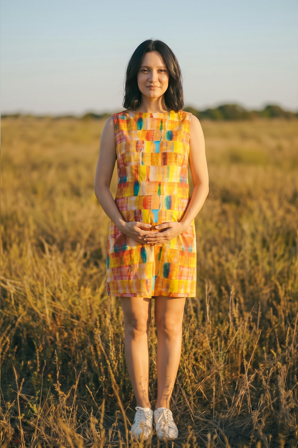A woman stands in a dry grass field at sunset wearing a sleeveless, knee-length hemp dress with a colorful patchwork pattern of orange, yellow, blue, and pink blocks, paired with white sneakers.