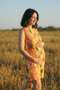 A pregnant woman stands in a sunlit grassy field wearing a sleeveless, knee-length hemp dress with a colorful abstract patchwork pattern in yellow, orange, pink, and blue.