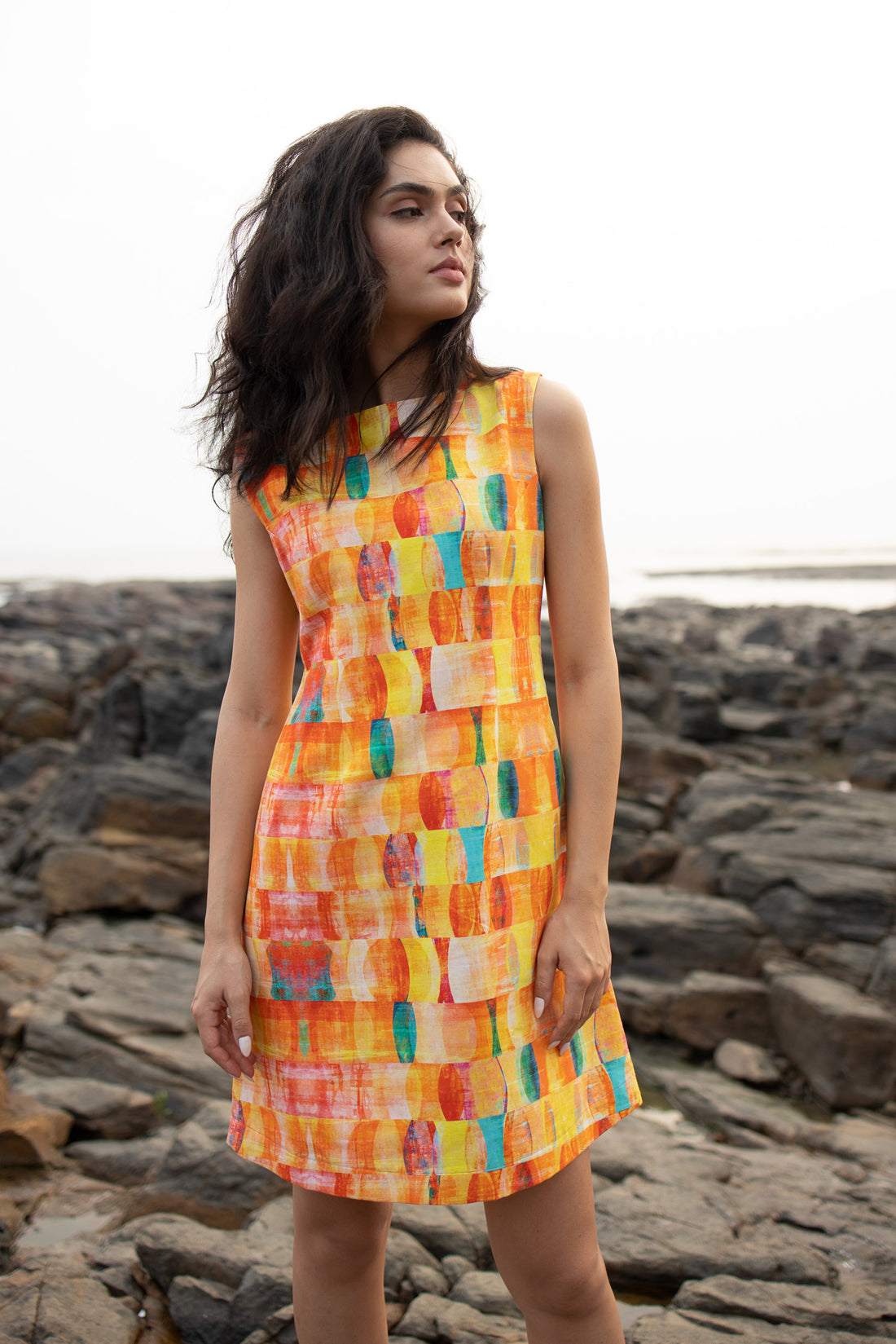 A woman wearing a sleeveless, knee-length multicolor hemp dress with a boat neckline, standing on a rocky beach.