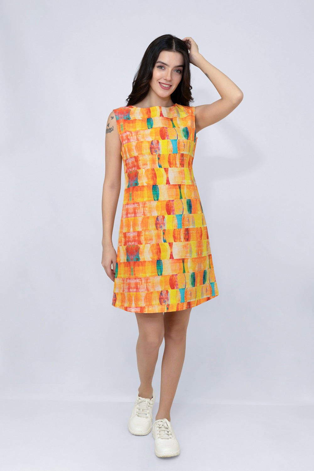 A woman modeling a sleeveless A-line hemp dress with a colorful abstract block pattern in orange, yellow, red, and blue, paired with white sneakers, against a light gray studio background.