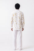 A man viewed from behind wearing a white long-sleeve button-up shirt with a scattered gold pattern and white trousers, standing against a plain white studio background.