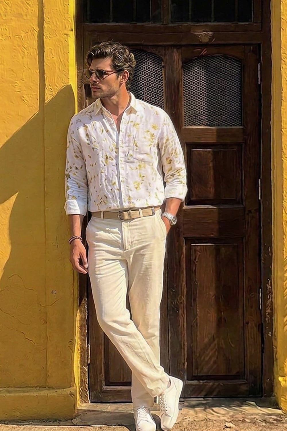 A man wearing a white long-sleeve button-up shirt with a yellow floral print, cream trousers, and white sneakers stands in front of a yellow wall and dark wooden door.