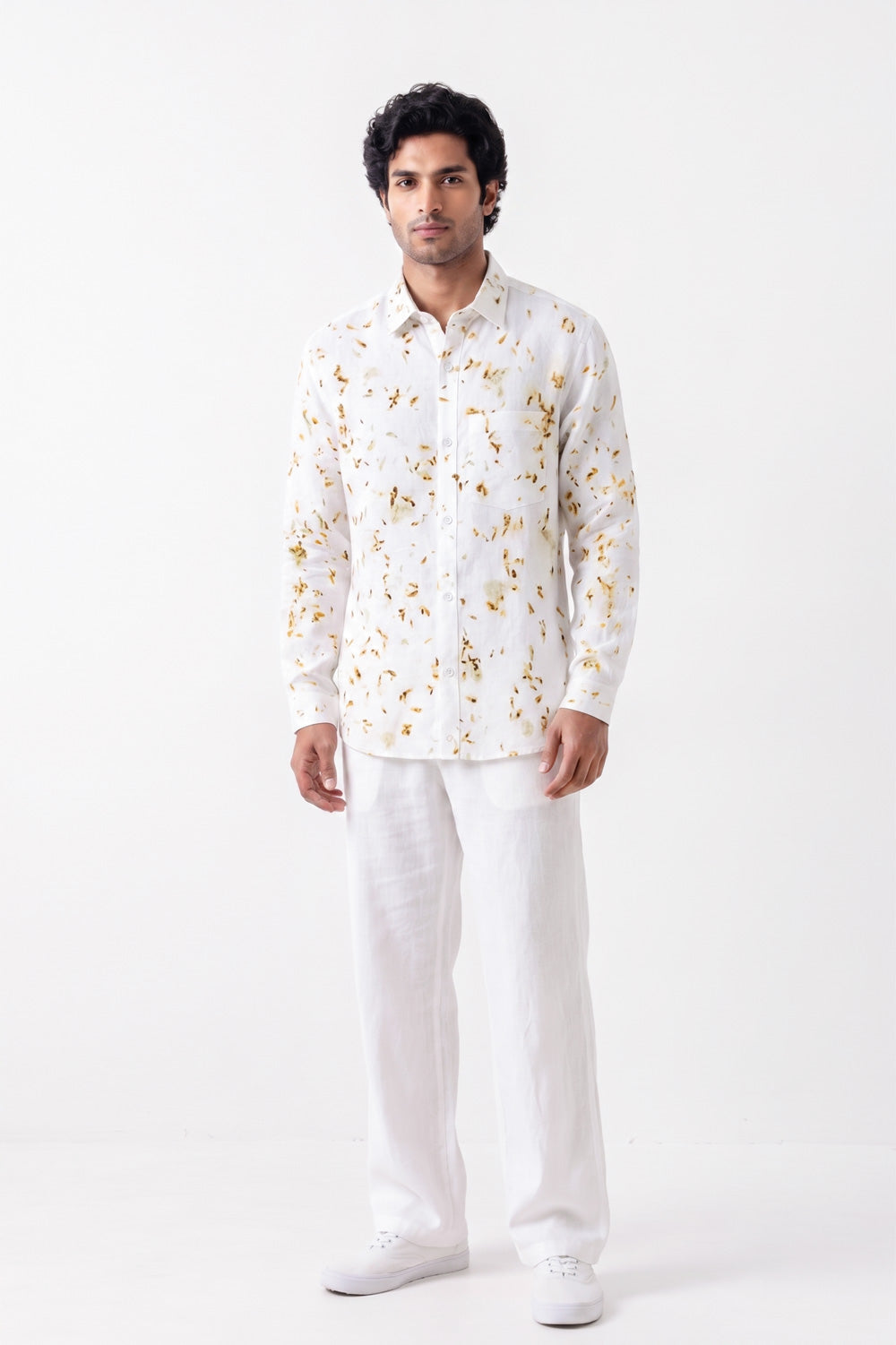 A man modeling a white long-sleeve button-up shirt with a delicate gold floral pattern, paired with white trousers and white sneakers, against a plain white studio background.