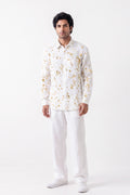 A man modeling a white long-sleeve button-up shirt with a delicate gold floral pattern, paired with white trousers and white sneakers, against a plain white studio background.