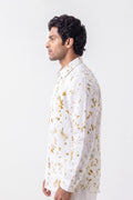 A man shown in profile wearing a white long-sleeve kurta with a gold floral pattern, photographed against a plain light gray studio background.