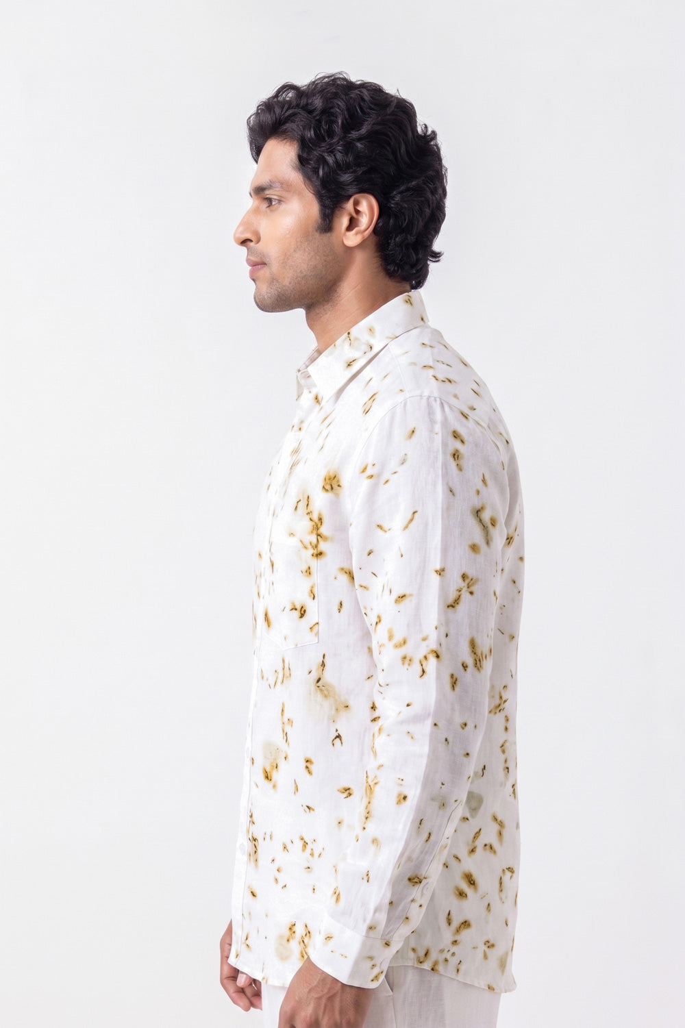 A man shown in profile wearing a white long-sleeve kurta with a gold floral pattern, photographed against a plain light gray studio background.